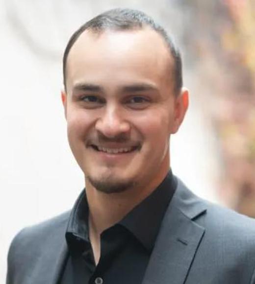 Headshot of a smiling man with short hair and goatee in dark suit, blurred outdoor background