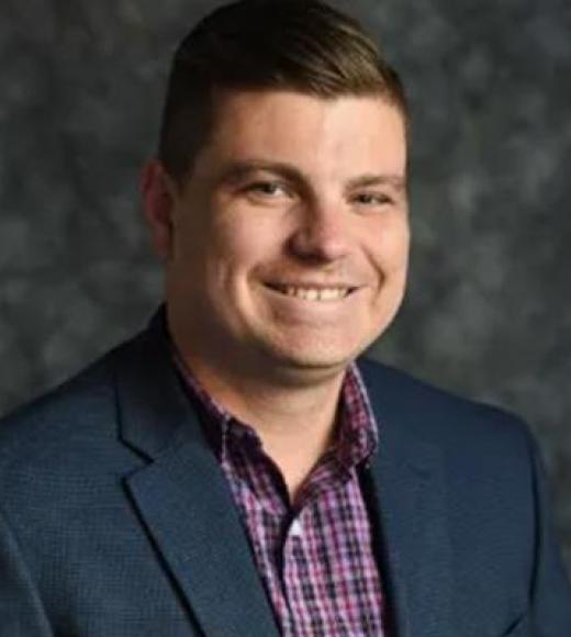 Smiling man in navy blazer and purple checked shirt, headshot on mottled gray background