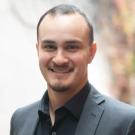Headshot of a smiling man with short hair and goatee in dark suit, blurred outdoor background