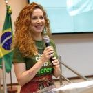 Smiling red-haired woman in green shirt holding microphone at podium, Brazilian flag behind