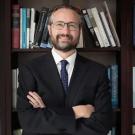 Smiling bearded man in dark suit and tie, arms crossed before bookshelf (photograph)
