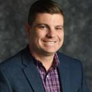 Smiling man in navy blazer and purple checked shirt, headshot on mottled gray background