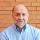 Portrait photo of older bald man with grey beard in blue shirt against brick wall
